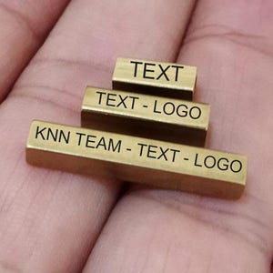 Può includere: Tre piccole barre metalliche rettangolari color oro impilate l'una sull'altra. Ogni barra ha un testo inciso: "TEXT", "TEXT - LOGO" e "KNN TEAM - TEXT - LOGO". Le barre poggiano su una superficie color carne.