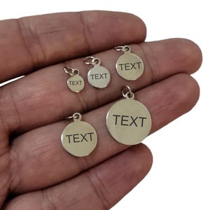 以下が含まれることがあります： 「TEXT」と刻まれた銀色の丸い金属タグが4つ。タグはサイズが異なり、手で持たれています。