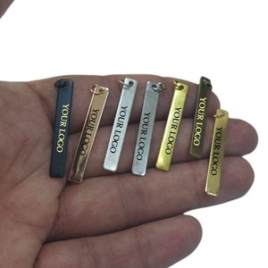 Puede incluir: Siete etiquetas metálicas diferentes con forma rectangular y esquinas redondeadas. Cada etiqueta tiene un agujero en la parte superior para sujetarse a un producto. Las etiquetas están disponibles en varios acabados, incluyendo negro, plata, oro y latón. Cada etiqueta tiene el texto "YOUR LOGO" impreso en letras doradas.