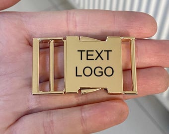 May include: A gold-colored metal buckle with the words "TEXT LOGO" printed in black. The buckle has a rectangular center with a sliding mechanism and two rectangular side pieces.