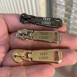 Może przedstawiać: Zbliżenie trzech metalowych suwaków w kolorze czarnym, srebrnym i złotym. Każdy suwak ma prostokątną płytkę z napisem "TEXT LOGO". Suwaki są przeznaczone do użytku w odzieży lub akcesoriach.
