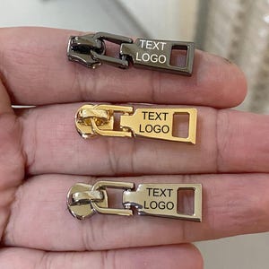 May include: Three metal zipper pulls in gunmetal, gold, and silver are displayed. Each pull features a rectangular design with the text "TEXT LOGO". The zipper pulls are held in a hand.