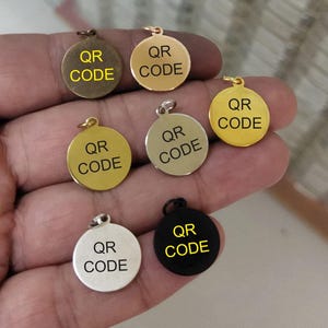 May include: A selection of circular metal charms in gold, silver, and black. Each charm has a small loop at the top and displays the text "QR CODE". These charms are designed for use as pendants or tags. The charms are in various metallic finishes.