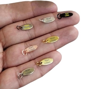 Op de afbeelding: Zes ovale metalen labels met springringen. Elk label heeft de tekst "TEXT LOGO" gegraveerd. De labels zijn in verschillende kleuren: zwart, zilver, goud, rosé goud en twee tinten messing.