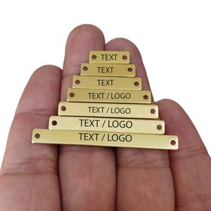 Può includere: Sette etichette in metallo dorate con bordi arrotondati e fori nella parte superiore. Le etichette sono disposte a forma di piramide. Le etichette sono incise con le parole "TEXT" e "TEXT/LOGO".