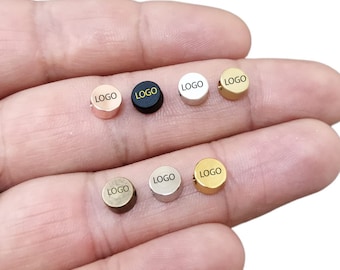 May include: Seven round metal charms with the word "LOGO" engraved on each. The charms are in different colours: rose gold, black, silver, gold, bronze, and two shades of grey.