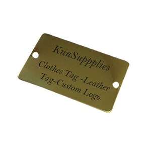 Op de afbeelding: Een gouden metalen label met de tekst "KnnSupplies Clothes Tag - Leather Tag - Custom Logo" erop gegraveerd.