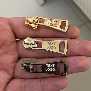 Puede incluir: Tres tiradores de cremallera metálicos en oro, plata y negro. Cada tirador tiene una placa rectangular con el texto "TEXT LOGO". Los tiradores se sostienen en una mano.
