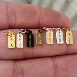 Può includere: Una collezione di piccoli ciondoli rettangolari in metallo di vari colori, tra cui oro, argento e nero. Ogni ciondolo ha un anello nella parte superiore e il testo "TEXT / LOGO" inciso sul davanti. Questi ciondoli sono probabilmente utilizzati per gioielli o progetti artigianali.
