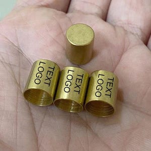 May include: A collection of small, cylindrical brass objects. Three of the cylinders have the word "TEXT" and "LOGO" printed on them. One solid cylinder is also present. The objects are displayed on a light-colored surface.