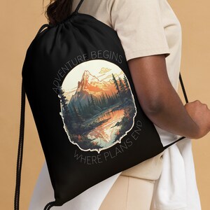 Può includere: Zaino nero con coulisse con un design circolare di montagna e lago. L'immagine presenta una catena montuosa, alberi e acqua, con il testo "ADVENTURE BEGINS WHERE PLANS END" in bianco.