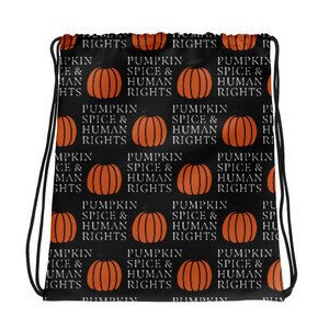 Peut inclure: Sac de sport noir avec un motif répété de citrouilles oranges et le texte "PUMPKIN SPICE & HUMAN RIGHTS" en blanc. Le sac a des cordons noirs pour la fermeture.