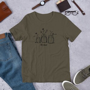 May include: Olive green t-shirt featuring a black line drawing of three mason jars with flowers. The text "Be kind" is printed below the jars. The shirt is made of a soft material.