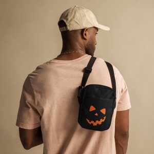 May include: Black crossbody bag with an orange jack-o'-lantern face. The bag has a black adjustable strap and a buckle closure. The bag is worn over the shoulder. The person in the image is wearing a light pink t-shirt and a beige baseball cap.
