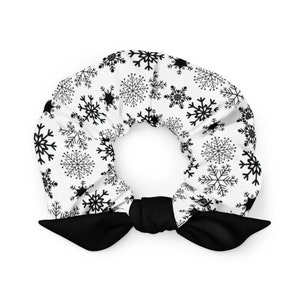 May include: A white scrunchie with a black snowflake pattern and a black bow. The scrunchie is a circular shape, perfect for holding hair. The snowflake design adds a festive touch to this hair accessory.