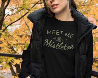 May include: Black long-sleeved top with the text "MEET ME at the Mistletoe" in a light green font. The top has a crew neck and is worn by a person wearing a black vest. The background features autumn foliage.
