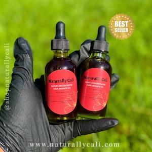 K&ouml;nnte beinhalten: Zwei Glasflaschen Naturally Cali Super Concentrated Hair Growth Oil mit schwarzen Tropfern und roten Etiketten. Die Flaschen werden in einer behandschuhten Hand vor einem gr&uuml;nen Hintergrund gehalten. Der Text "BEST SELLER" steht in einem goldenen Stern.