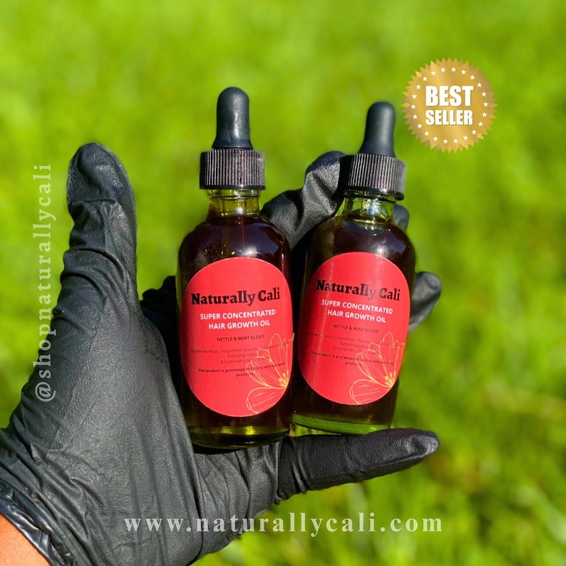 K&ouml;nnte beinhalten: Zwei Glasflaschen Naturally Cali Super Concentrated Hair Growth Oil mit schwarzen Tropfern und roten Etiketten. Die Flaschen werden in einer behandschuhten Hand vor einem gr&uuml;nen Hintergrund gehalten. Der Text "BEST SELLER" steht in einem goldenen Stern.