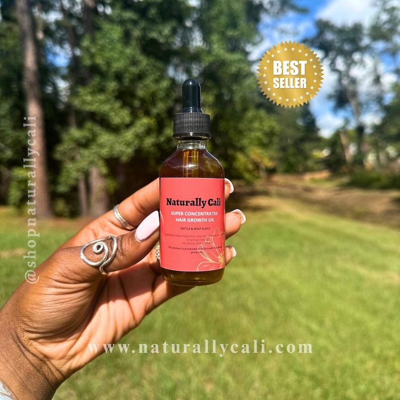 K&ouml;nnte beinhalten: Eine braune Glasflasche mit Naturally Cali Super Concentrated Hair Growth Oil, gehalten in einer Hand. Die Flasche hat eine schwarze Pipette und ein rotes Etikett mit wei&szlig;em Text. Ein goldener "Best Seller"-Aufkleber ist sichtbar. Der Hintergrund ist eine gr&uuml;ne Wiese und B&auml;ume.