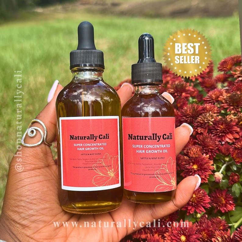 K&ouml;nnte beinhalten: Zwei Glasflaschen Naturally Cali Super Concentrated Hair Growth Oil mit schwarzen Tropfern und roten Etiketten. Die Etiketten enthalten den Text "Nettle & Mint Blend" und "Best Seller" in einem goldenen Stern. Die Flaschen werden in einer Hand gehalten.