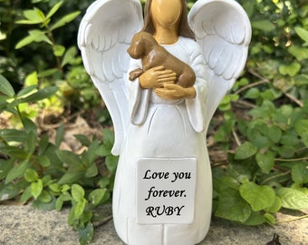 May include: White angel figurine holding a brown puppy. The angel has wings and is standing on a gray surface. The figurine has a plaque that reads "Love you forever. RUBY".