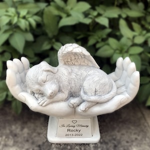 May include: White ceramic memorial statue of a sleeping dog with angel wings resting in cupped hands. The base has the inscription "In Loving Memory Rocky 2013-2022".