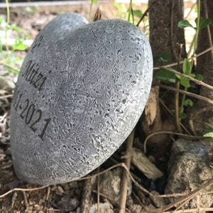 May include: A grey heart-shaped stone with the inscription "Mitzi 11-1-2021" on it. The stone is sitting on a bed of brown leaves and twigs.