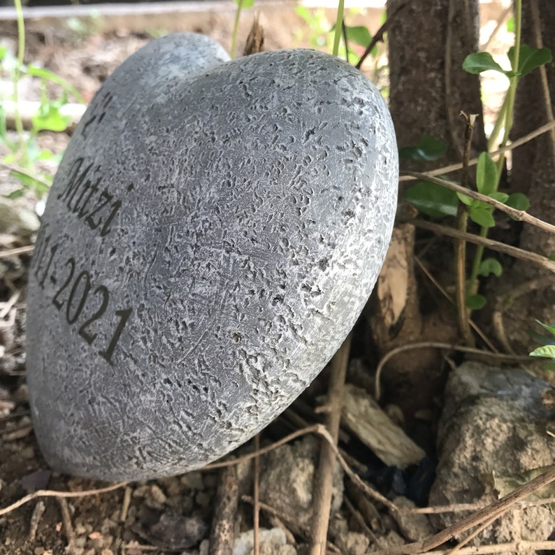 May include: A grey heart-shaped stone with the inscription "Mitzi 11-1-2021" on it. The stone is sitting on a bed of brown leaves and twigs.