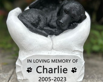 May include: White ceramic memorial statue of a black dog with wings, sleeping in a pair of hands. The statue has a plaque with the text "In loving memory of Charlie 2005-2023" and paw prints.