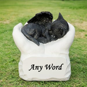 Può includere: Scultura commemorativa in ceramica bianca di un gatto nero con le ali che riposa in un paio di mani. La scultura ha uno spazio vuoto per l'iscrizione con le parole "Any Word" scritte su di essa.