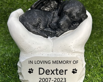 May include: White ceramic memorial figurine of a black puppy with angel wings sleeping in a pair of hands. The base has the inscription "In loving memory of Dexter 2007-2023" with paw prints.
