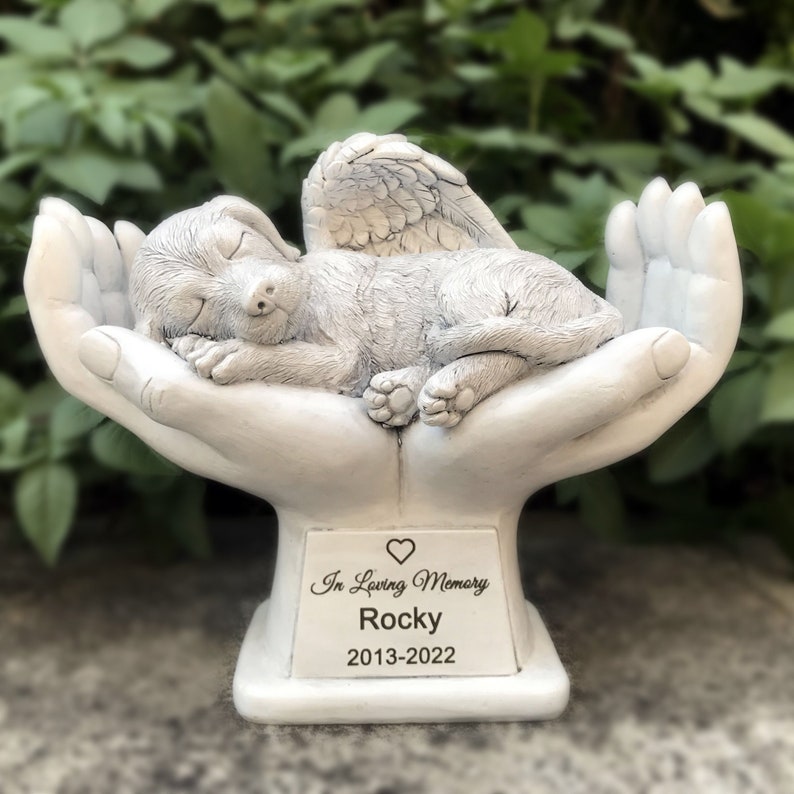 May include: White ceramic memorial statue of a sleeping dog with angel wings resting in cupped hands. The base has a plaque with the inscription "In Loving Memory Rocky 2013-2022".