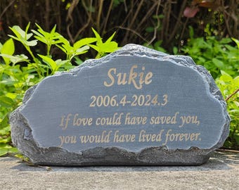 May include: A gray stone memorial plaque with the name "Sukie", dates, and the inscription "If love could have saved you, you would have lived forever." The text is in a gold color. The plaque has a natural, rough edge.