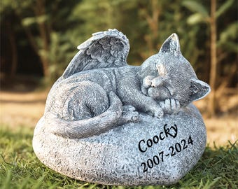 May include: A gray stone sculpture of a sleeping cat with angel wings resting on a heart-shaped stone. The stone has the inscription "Coocky 2007-2024" on it.