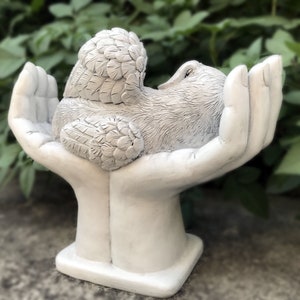 May include: A white ceramic sculpture of two hands holding a small winged bird with its wings folded. The bird is resting in the palms of the hands, which are raised up.