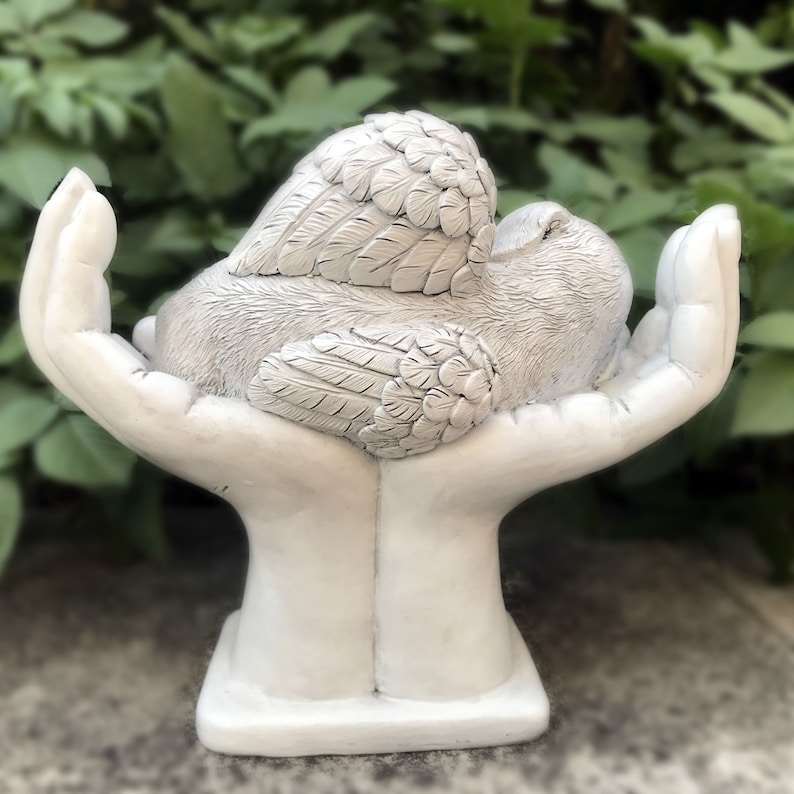 May include: White ceramic sculpture of two hands holding a bird with wings. The bird is sleeping with its head resting on its wings. The sculpture is a memorial or remembrance piece.