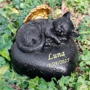 Op de afbeelding: Zwarte kat gedenkbeeldje met gouden vleugels, slaapt op een hartvormige steen met de inscriptie "Luna 3/29/2025".