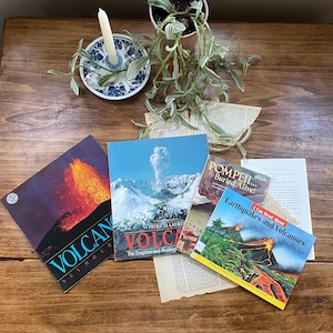 Può includere: Una collezione di libri sui vulcani e i terremoti, con titoli come "Volcan" e "Earthquakes and Volcanoes". I libri sono disposti su una superficie di legno, insieme a una piccola pianta in vaso e un portacandele.