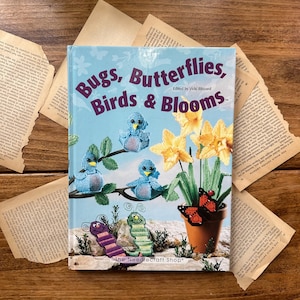May include: A book titled "Bugs, Butterflies, Birds & Blooms" with a blue cover featuring illustrations of crocheted birds, butterflies, and flowers. The book is surrounded by torn pages of an old book, set on a wooden surface.