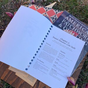 Pode incluir: Livro aberto com fundo branco e o t&iacute;tulo "Sweet Dreams Crib Quilt" na p&aacute;gina. O livro est&aacute; sobre uma superf&iacute;cie de madeira com grama verde e uma flor rosa no fundo.
