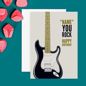 May include: Greeting card featuring a black and white electric guitar illustration. The card reads "*NAME* YOU ROCK HAPPY BIRTHDAY" in black and gold. Pink guitar picks are scattered on a teal surface.