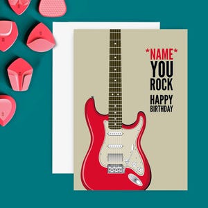 May include: A birthday card featuring a red electric guitar illustration with the text "*NAME* YOU ROCK HAPPY BIRTHDAY". The card is on a teal surface with pink guitar picks.