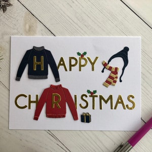 May include: A white greeting card with the words "HAPPY CHRISTMAS" in gold lettering. The card features navy and red sweater cutouts with gold letters, a black hat, and a red and gold scarf. A small gift box and holly accents are also present.