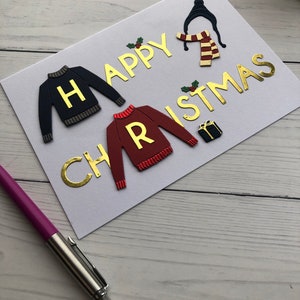 May include: A handmade Christmas card with gold lettering that reads "HAPPY CHRISTMAS". The card features sweater and scarf designs, along with a small gift box and holly accents. A purple pen rests on the wooden surface.