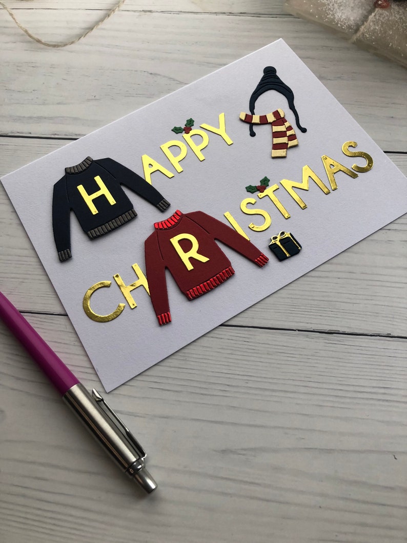May include: A handmade Christmas card with gold lettering that reads "HAPPY CHRISTMAS". The card features sweater and scarf designs, along with a small gift box and holly accents. A purple pen rests on the wooden surface.
