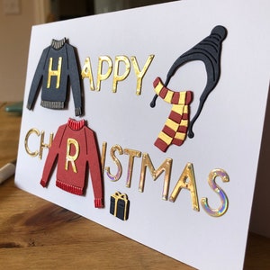 May include: A handmade Christmas card with the words "HAPPY CHRISTMAS" in gold lettering. The card features cut-out sweater and scarf designs, with the letters "H" and "R" on the sweaters. A small gift box is also included.