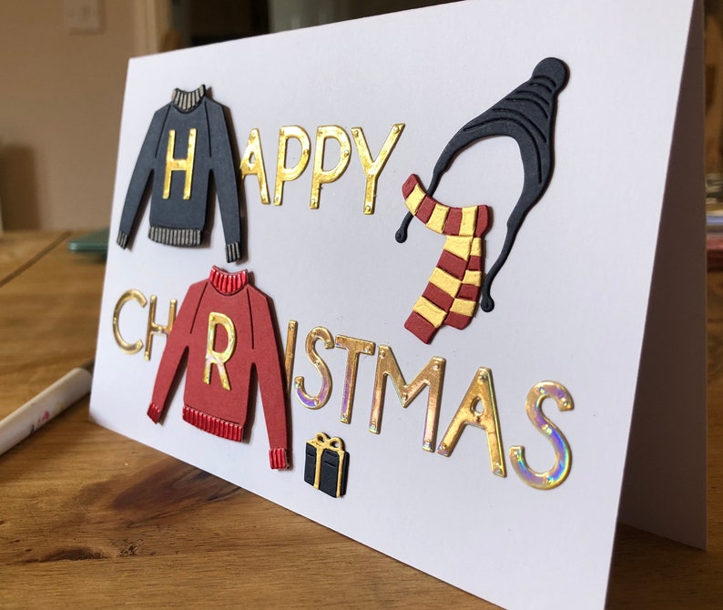 May include: A handmade Christmas card with the words "HAPPY CHRISTMAS" in gold lettering. The card features cut-out sweater and scarf designs, with the letters "H" and "R" on the sweaters. A small gift box is also included.
