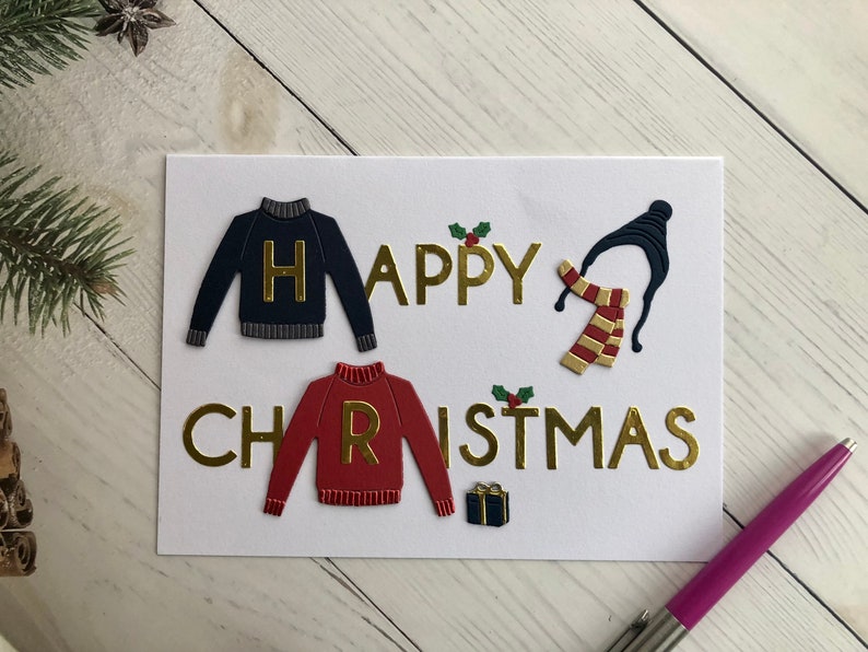May include: A white greeting card with the words "HAPPY CHRISTMAS" in gold lettering. The card features navy and red sweater cutouts with gold letters, a black hat, and a red and gold scarf. A small gift box and holly accents are also present.