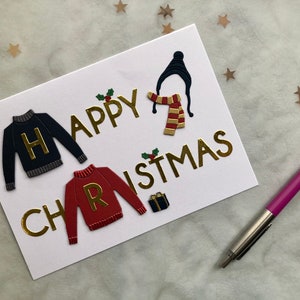 May include: A white greeting card with gold lettering that reads "Happy Christmas." The card features embossed images of a black sweater with a gold "H," a red sweater with a gold "R," a hat, scarf, and a small gift box.