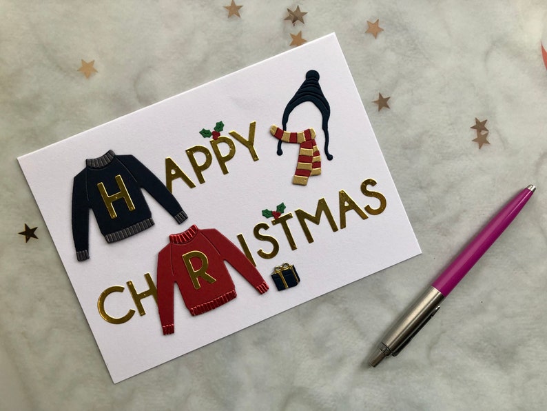 May include: A white greeting card with gold lettering that reads "Happy Christmas." The card features embossed images of a black sweater with a gold "H," a red sweater with a gold "R," a hat, scarf, and a small gift box.